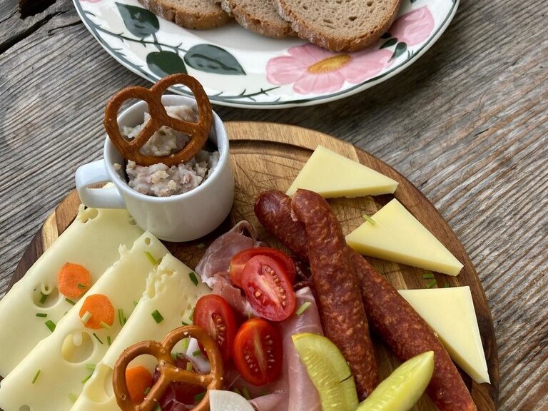 Eine gemischte Platte mit Wurst, Käse und anderen Köstlichkeiten steht neben einem Teller mit Brot auf einem Holztisch.